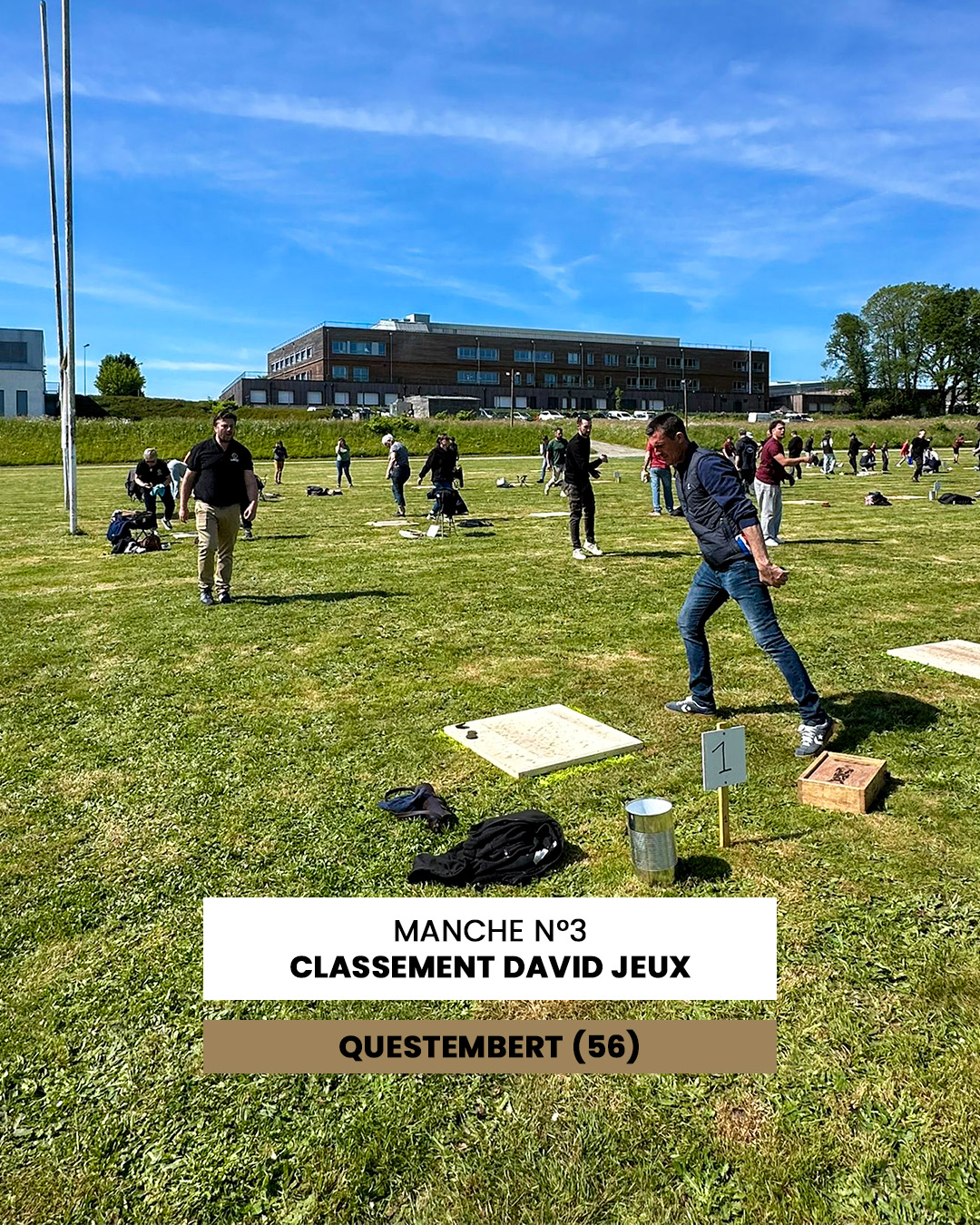 Outdoor field with people participating in a games competition; markers and boards are on the grass, sunny day. (Signage: 'MANCHE N°3 CLASSEMENT DAVID JEUX QUESTEMBERT (56)')