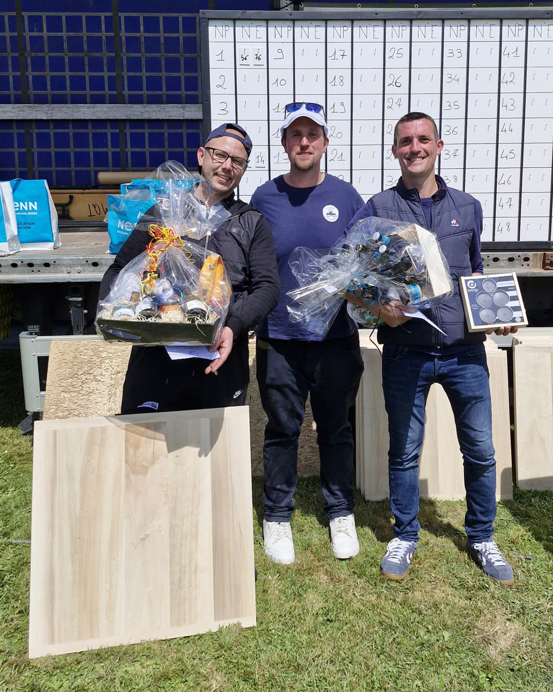 Three men stand outdoors, each holding gift baskets, posing for a prize photo with a numbered scoreboard behind them.