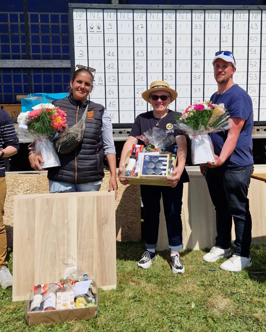 Three winners pose outdoors with bouquets and prize baskets in front of a large whiteboard-style scoreboard, on grass under bright sun.