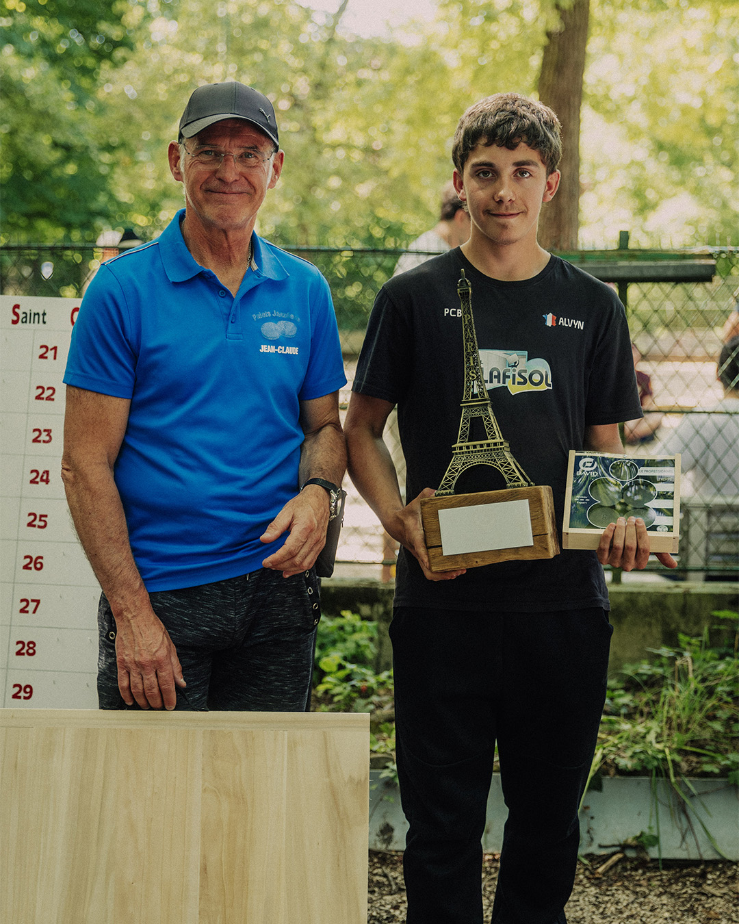 Two men posing outdoors after a trophy ceremony: an older man in a blue polo shirt and cap standing beside a young man holding a tall Eiffel Tower trophy and a boxed prize.