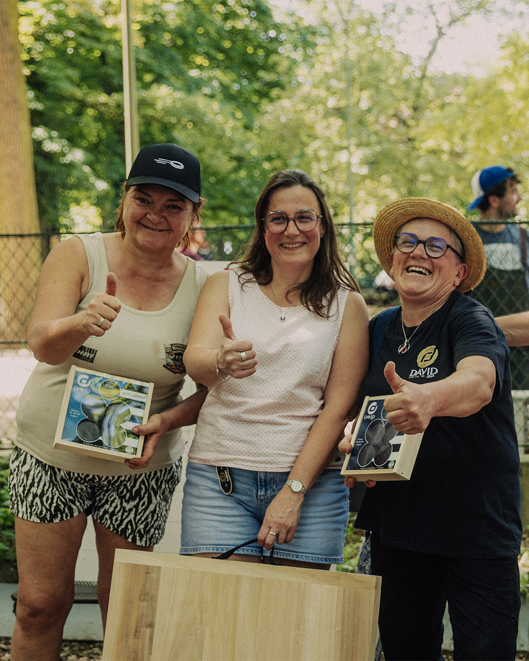 Three smiling women outdoors, giving thumbs up; two hold product boxes while a wooden board rests in front.