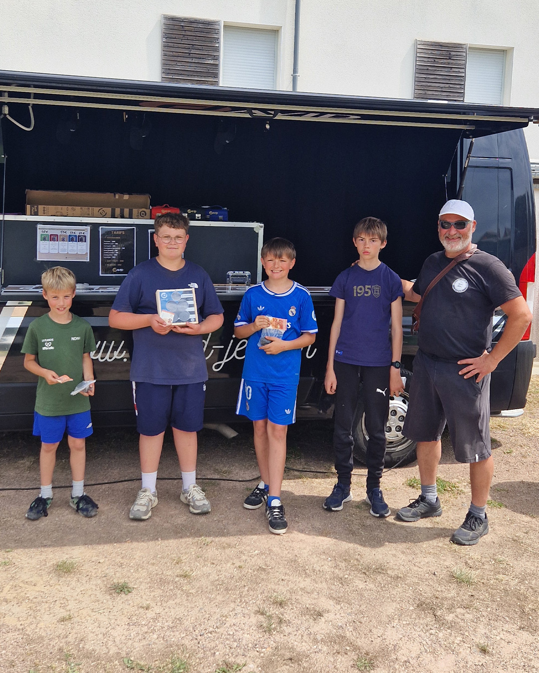 Group of five boys and an adult man posing in front of a black service truck with its back open, holding awards and prizes outdoors.
