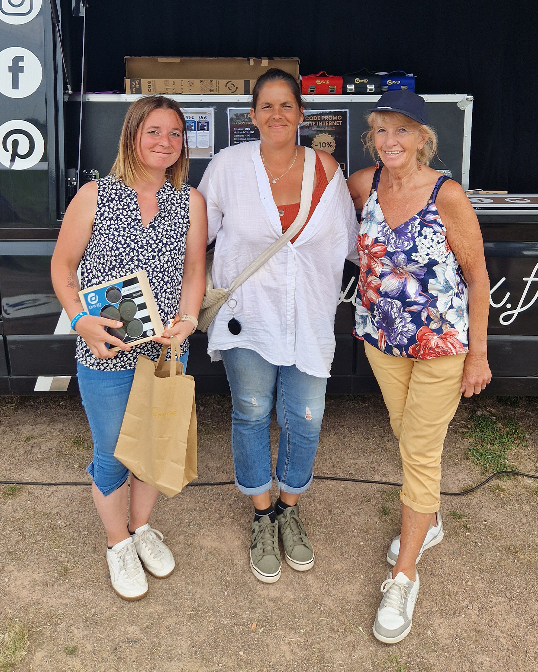 Three women pose in front of a black event trailer; left woman wears a sleeveless floral top and blue jeans, holding a paper bag and a boxed item; middle woman in a white blouse with a red top underneath and a crossbody bag; right woman in a floral sleeveless top and beige capri pants.